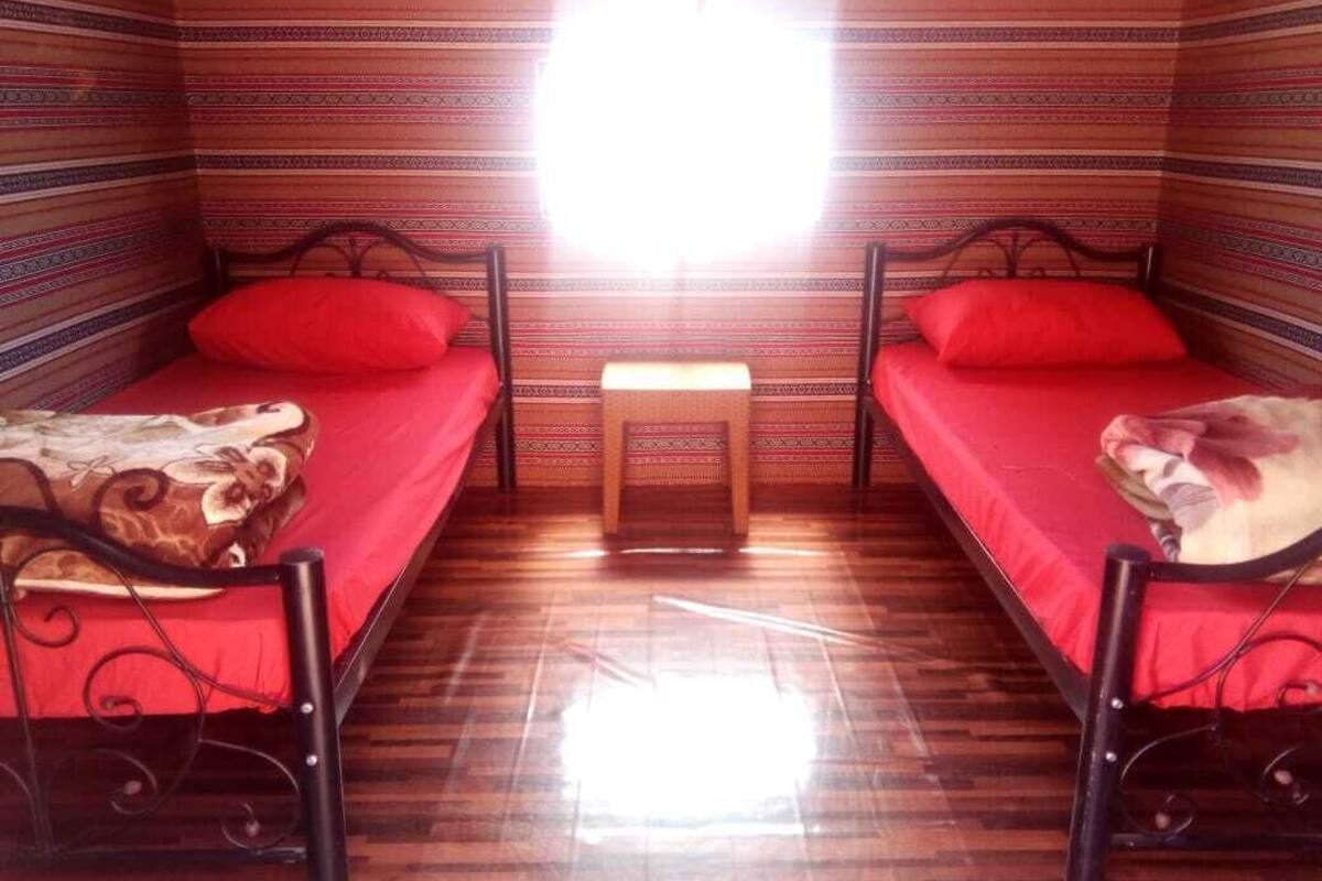 A room with two single beds, each with a red mattress and matching pillows. There is a small wooden stool in the center. The walls have a striped pattern, and the floor is wooden.