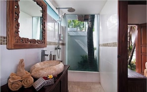 Un baño con un espejo grande, un mueble de tocador de madera, un lavabo de piedra y una ducha con paredes de vidrio que ofrece vistas a palmeras.