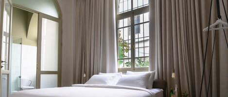 Ein Hotelzimmer mit einem großen Bett, einem Deckenventilator, einem Fenster mit Vorhängen und einem kleinen Holfnachtkästchen mit einer Flasche und einer Pflanze.