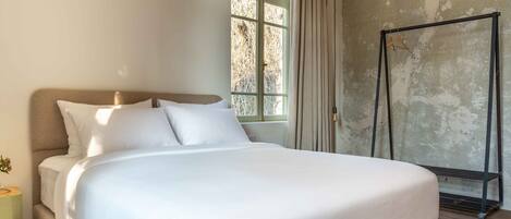 Una cama con sábanas blancas, una ventana con cortinas y un perchero metálico contra una pared con textura.