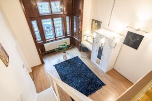 Huoneessa on puulattia, sinivärinen kuvioitu matto, patteri, ruukussa oleva kasvi, työpöytä tuolilla, kattokruunu ja seinälle ripustettu taideteos.