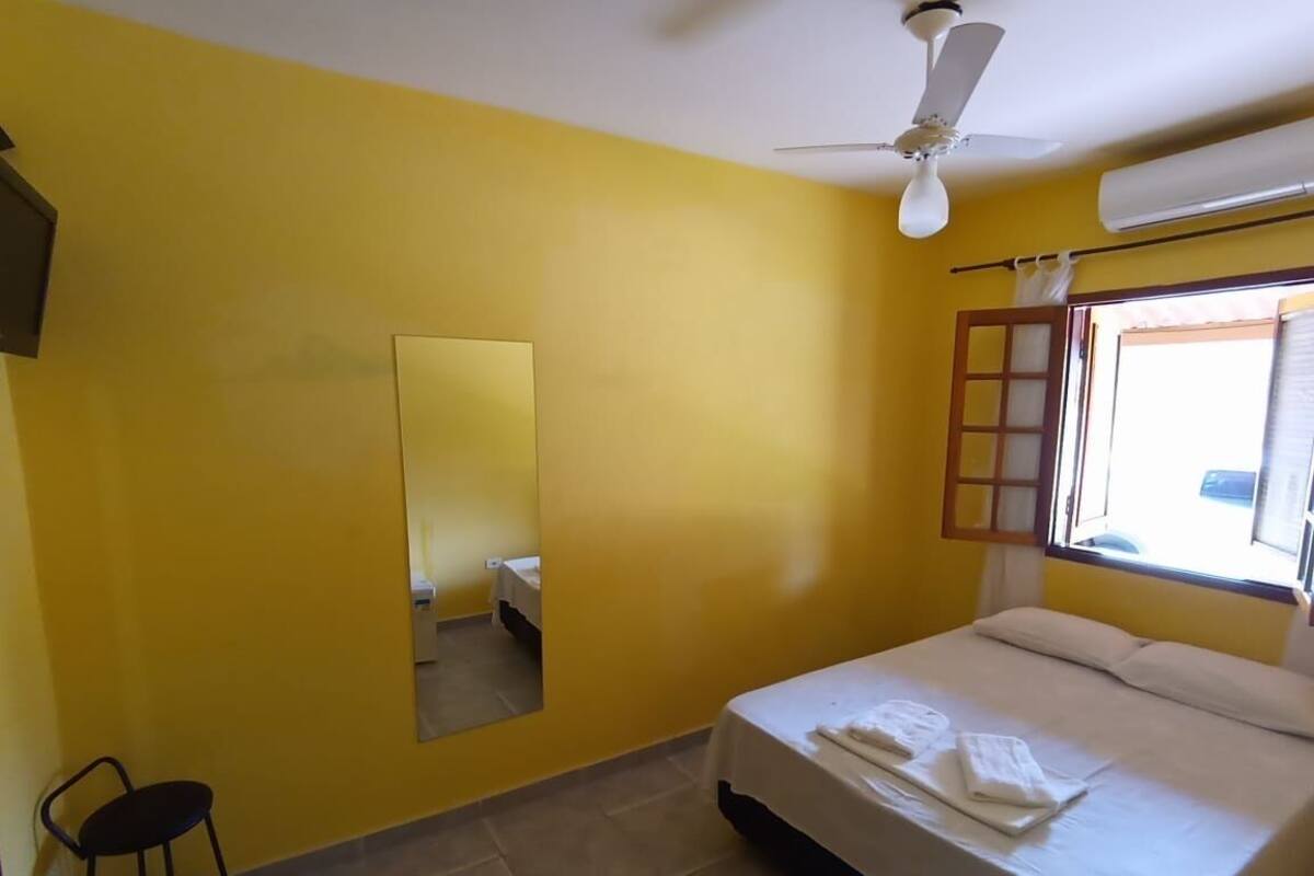 Una habitación con paredes amarillas, una cama con ropa blanca, un ventilador de techo, una ventana con marco de madera y un taburete pequeño.