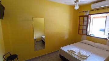 Quarto com paredes amarelas, cama com roupa de cama branca, ventilador de teto, janela com moldura de madeira e um pequeno banquinho.