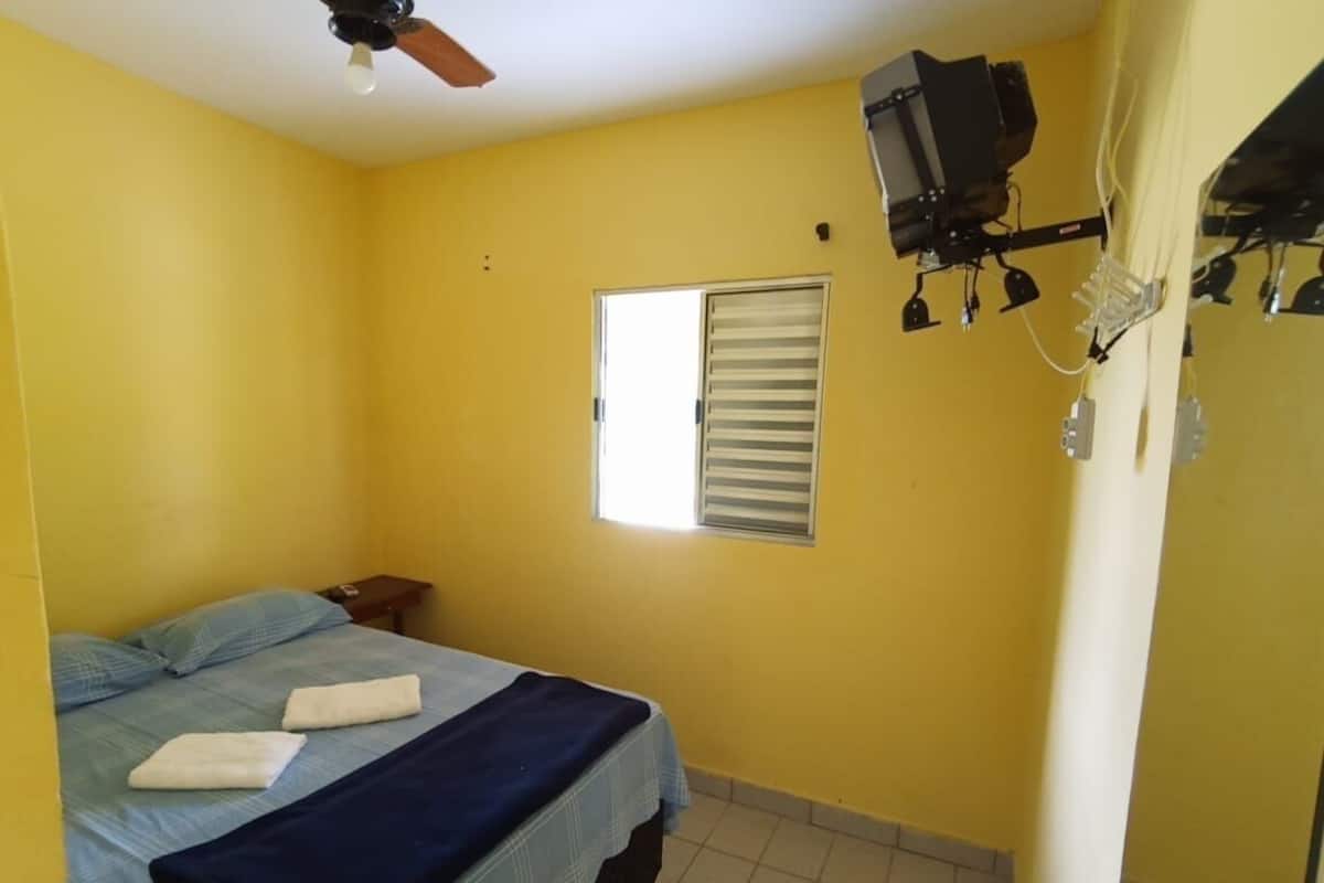 Una habitación con una cama, ventilador de techo, ventana con persianas y televisor montado en la pared.