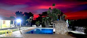 Area piscina notturna con struttura in pietra, poltrone da relax bianche e un'amaca.