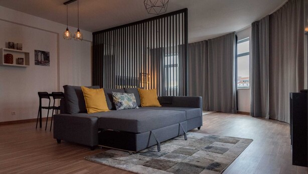 A modern living room with a grey sofa, yellow cushions, a patterned rug, and a black TV stand. - Round loft (azibo) (Macedo de Cavaleiros)