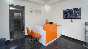 A reception area with a modern design, featuring a reception desk, three clocks, and a mounted television displaying multiple camera feeds. - PLLAZIO Residency (Gurugram)