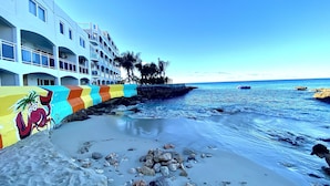 A beachfront hotel with a colorful mural, a rocky shoreline, and a clear blue sky.