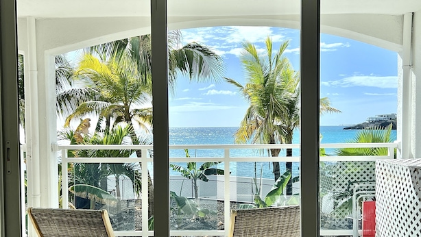 A balcony with two wicker chairs, a white lattice fence, and a view of the sea and palm trees.