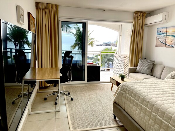 Quarto de hotel com cama, escrivaninha, cadeira, janela com cortinas e vista para o oceano.