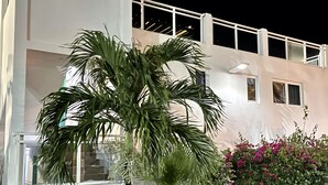 A multi-story building with balconies and a garden area featuring palm trees and flowering plants.