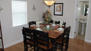 Une salle à manger avec une table en bois dressée pour six personnes, entourée de chaises noires. Un lustre est suspendu au-dessus de la table, et une fenêtre avec des stores se trouve à gauche.