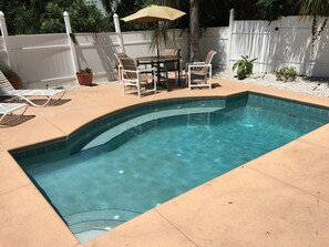 A swimming pool with a curved edge, surrounded by a patio area with a table and chairs, and a white fence in the background.