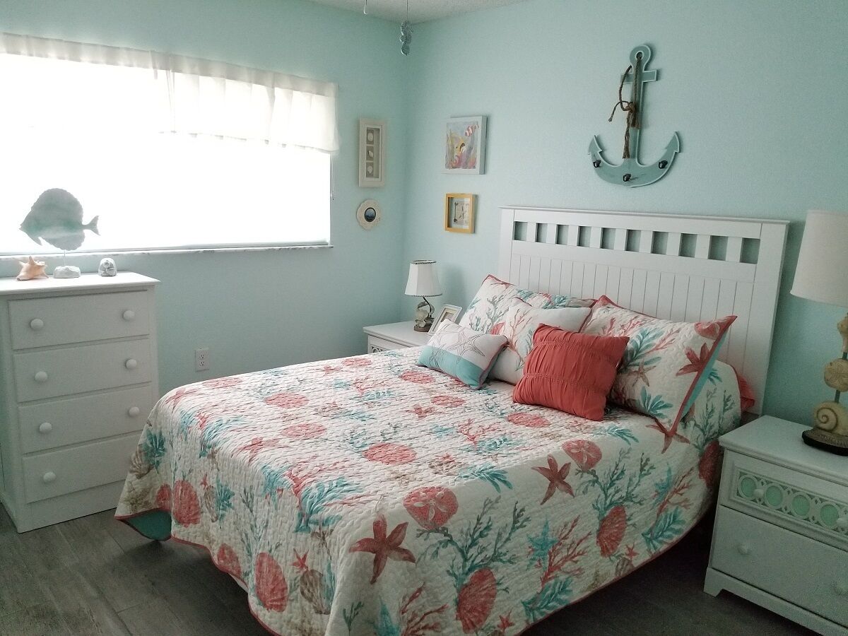 Un dormitorio con colcha estampada, cómoda blanca, mesita de noche con lámpara y decoración náutica en la pared.