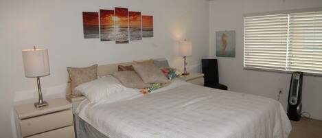 A bedroom with a bed, two bedside tables, a nightstand, a wall-mounted lamp, a window with blinds, and three wall art pieces.