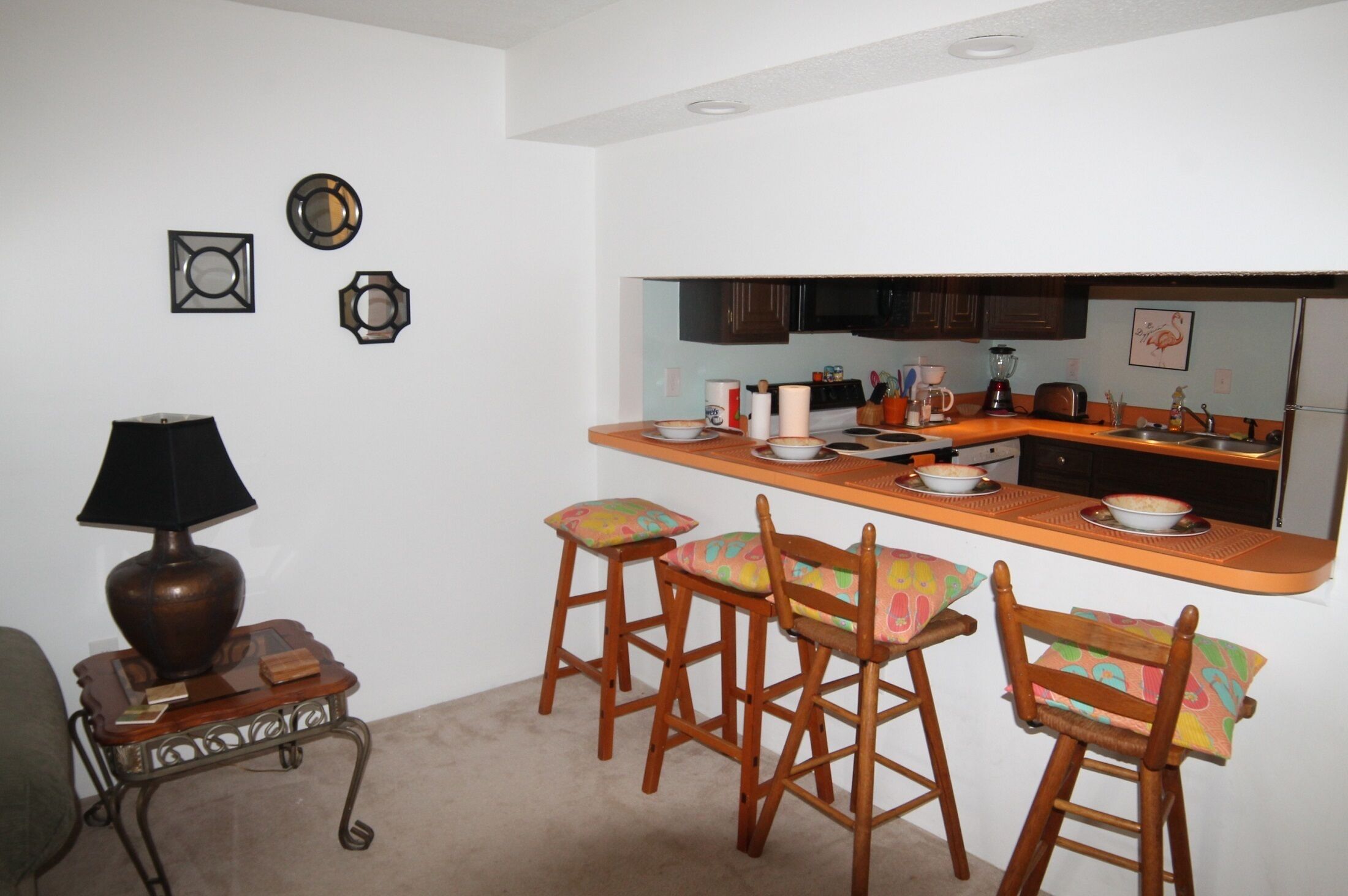 A compact kitchen with a bar area, wooden chairs, and a small side table with a lamp.