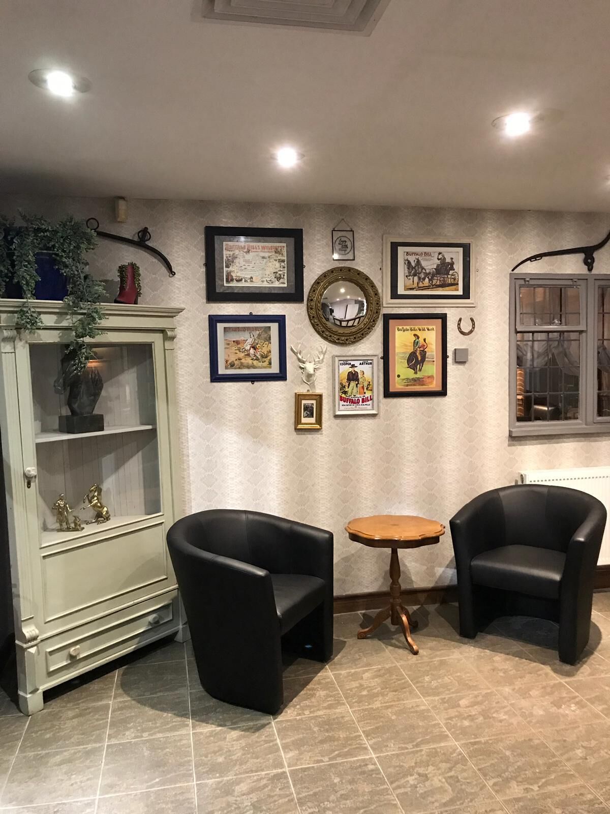 Una habitación con dos sillones de cuero negro, una mesita redonda pequeña, un aparador verde con objetos decorativos, y una pared decorada con cuadros y un espejo.