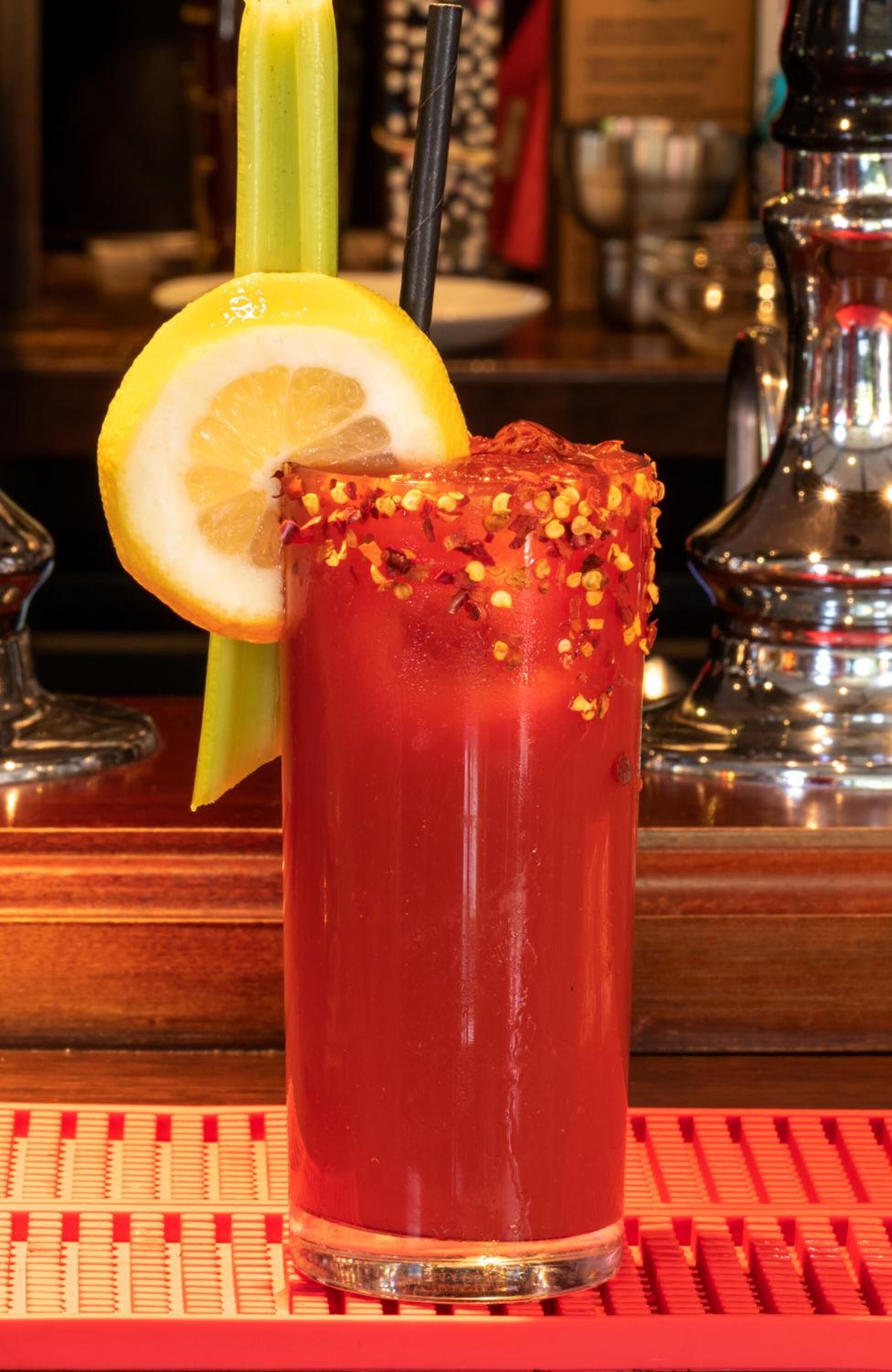 A tall glass of Bloody Mary with a lemon slice and celery stalk, garnished with spices, on a bar counter.