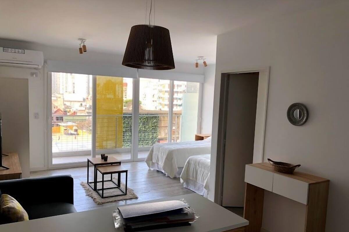 A modern, well-lit room with a large window, a bed, a dining table, and a small side table.