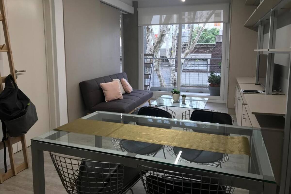 A modern living room with a glass dining table, a sofa, and a kitchen area.