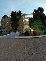 Entrada de um hotel com um caminho de paralelepípedos, cercada por árvores e arbustos.