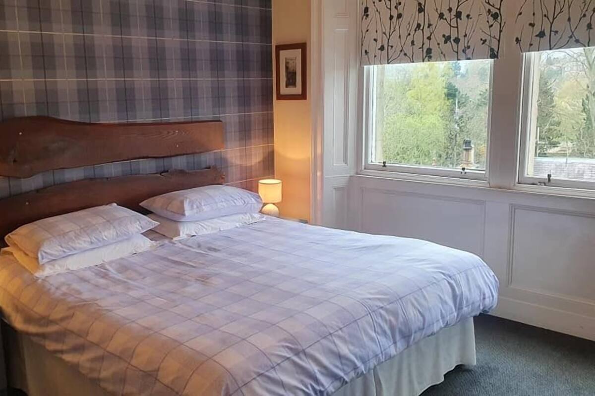 A bedroom with a large bed, two bedside tables, a headboard, and a window with a view of greenery.