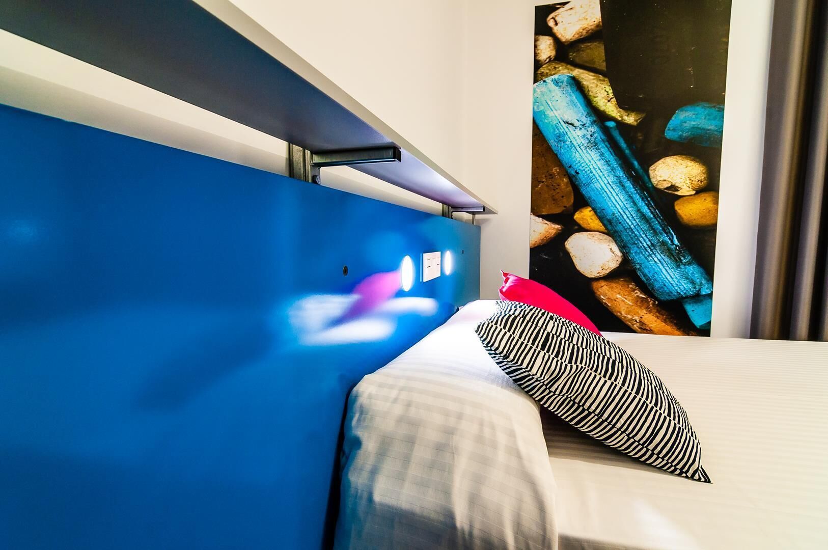 Habitació d'hotel amb un llit, una paret blava, una finestra que dona a unes roques i un almoiner vermell.