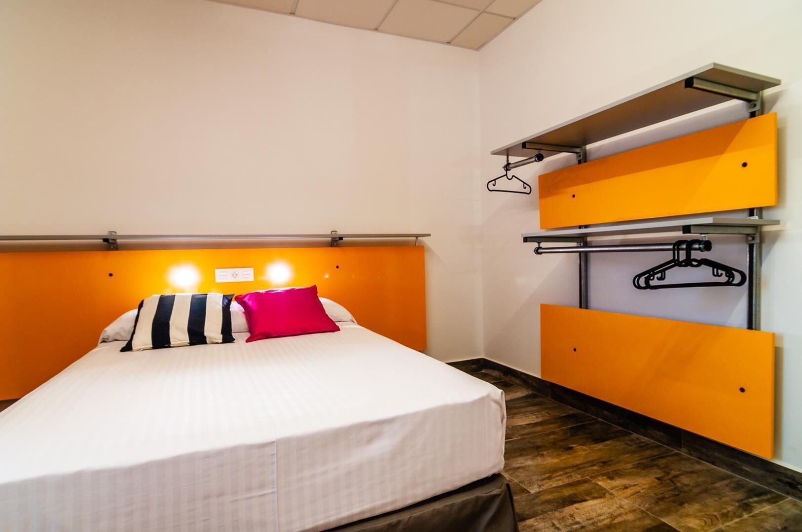 A hotel room with a bed, a headboard with lights, a wardrobe with a hanger, and a wooden floor.