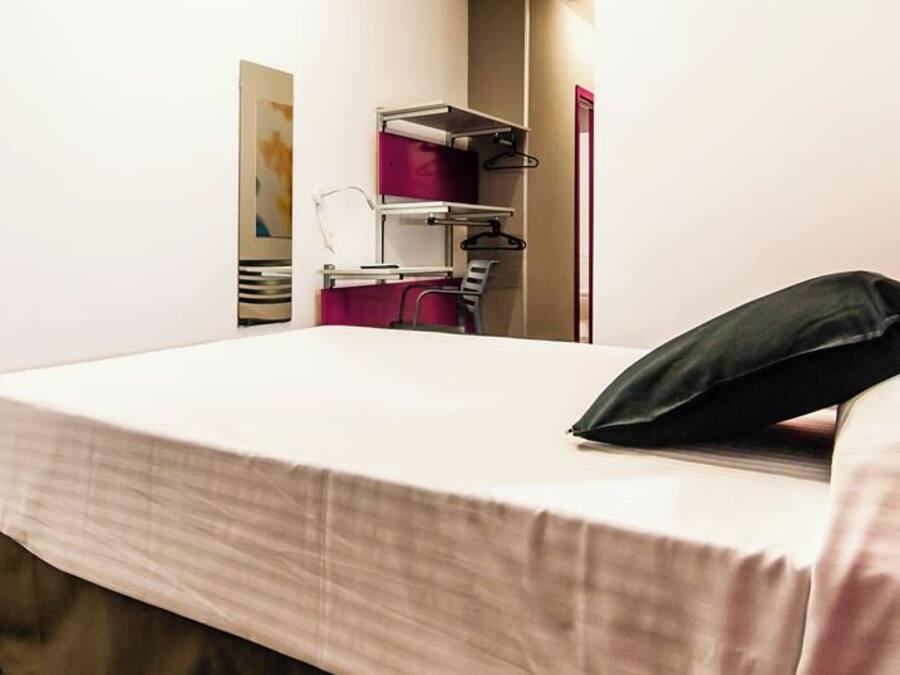 A hotel room with a bed, a flat-screen TV mounted on the wall, a small table with a chair, and a shelf with hanging items.