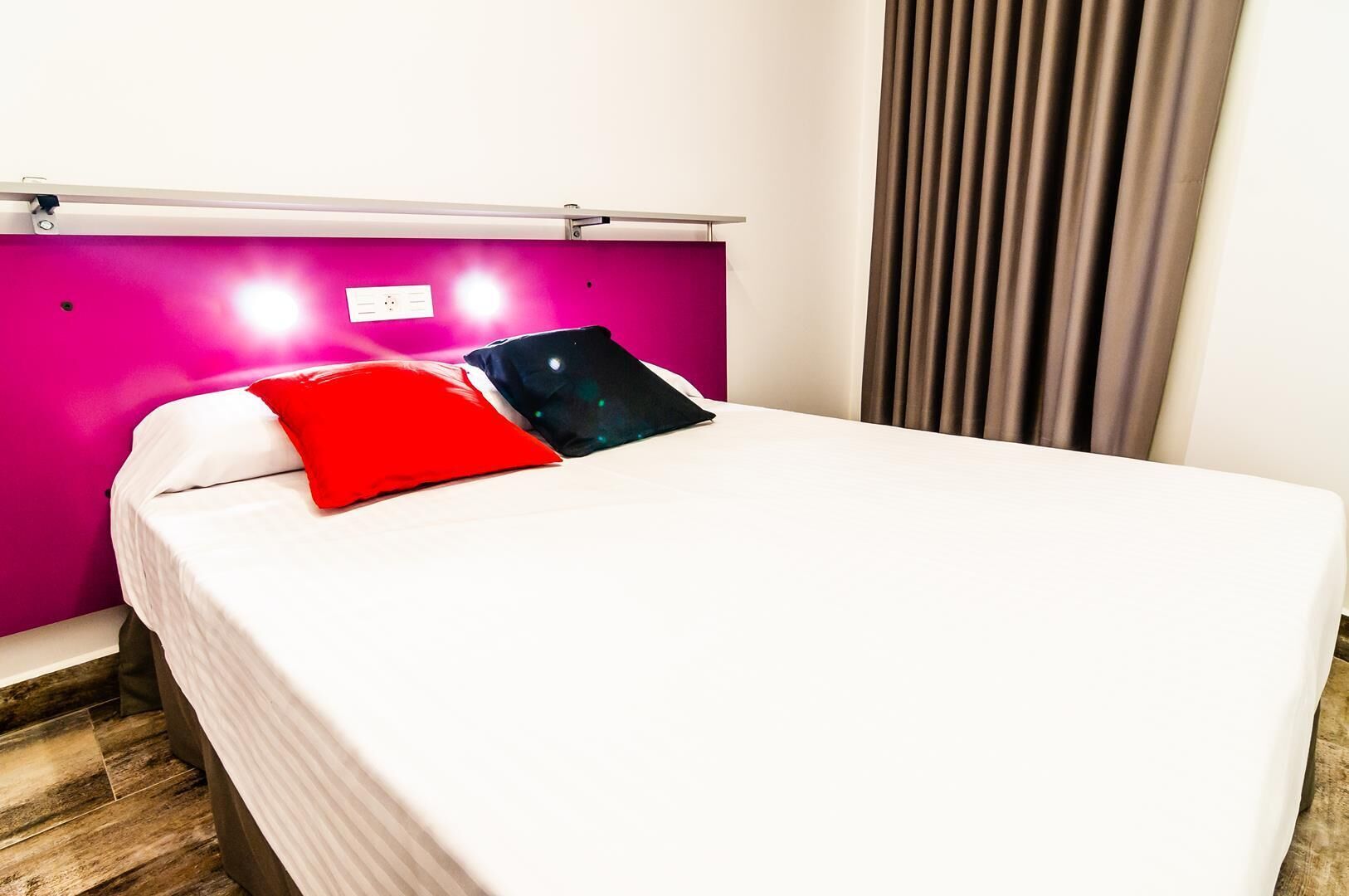 A hotel room with a bed, a bright pink wall, and a window with curtains.