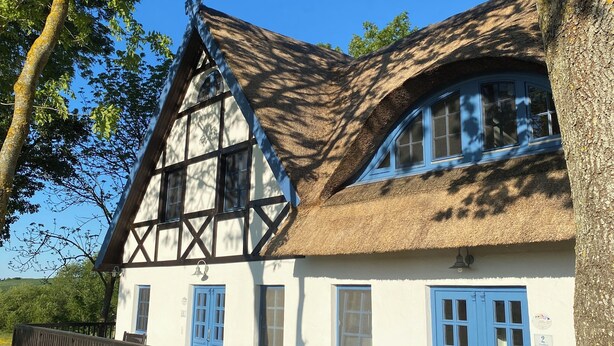 Une maison traditionnelle à colombages, avec un toit de chaume, des portes bleues et une terrasse en bois.