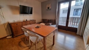 A room with a wooden dining table, two white chairs, a wooden cabinet, a flat-screen TV mounted on the wall, a sofa with patterned cushions, and a sliding glass door leading to an outdoor area.