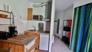 Eine kleine Küche mit Mikrowelle, Toaster, Spüle und kleinem Kühlschrank. Ein Holztisch mit Schneidebrett und Geschirr, sowie ein Stuhl sind ebenfalls zu sehen. Außerdem ist ein Regal mit Schüssel und Korb erkennbar.