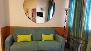 Ein grünes Sofa mit zwei gelben Kissen, ein runder Spiegel an der Wand, eine Stehlampe und ein gestreifter Vorhang.