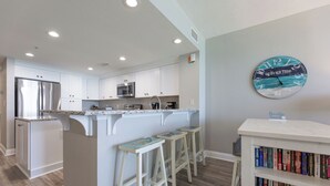 Una cocina moderna con armarios blancos, un refrigerador de acero inoxidable, un microondas y una cafetera. Hay una isla de madera con taburetes y una estantería con libros.