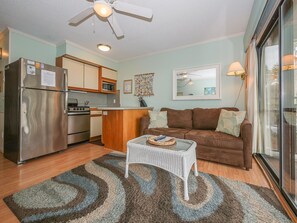 Un espacio habitable compacto con kitchenette, un sofá marrón, una mesita blanca y un ventilador de techo.