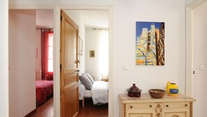 A hallway with a wooden door leading to a bedroom with a red bedspread. A painting of a tree is on the wall, and a cabinet with kitchenware is visible. - L'escalivade Chambres d'hôtes (Ceret)