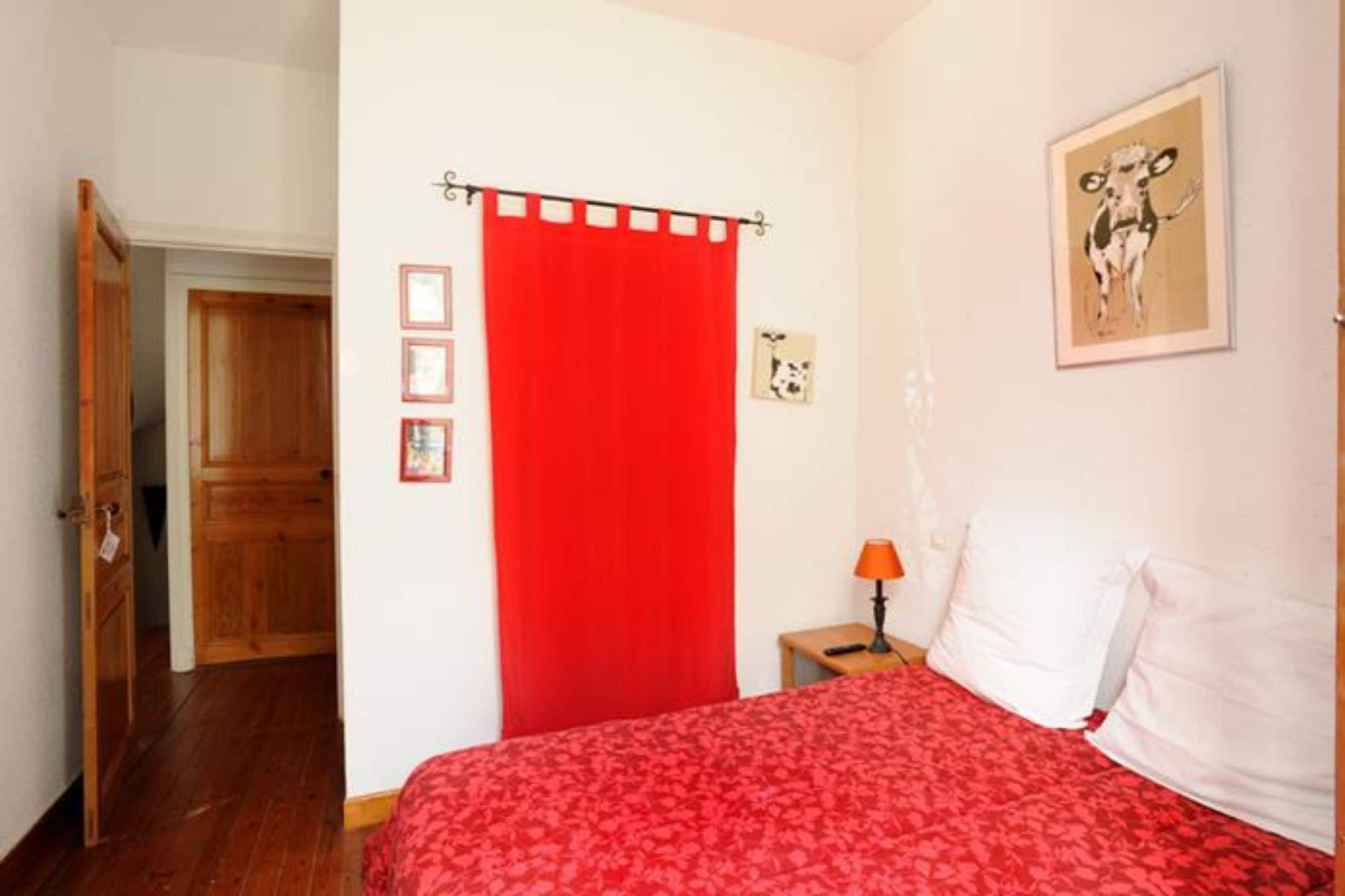 Een slaapkamer met een rode sprei, een houten deur, een rood gordijn, een klein nachtkastje met een lamp en een lijst met een foto aan de muur.