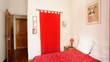 Een slaapkamer met een rode sprei, een houten deur, een rood gordijn, een klein nachtkastje met een lamp en een lijst met een foto aan de muur.