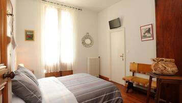 A bedroom with a bed, a window with curtains, a radiator, a television on the wall, and a wooden bench.