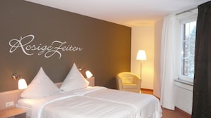 Habitació d'hotel amb un llit, dues làmpades de nit, una cadira, una finestra amb cortines i una paret amb la inscripció "Rosige Zeiten".