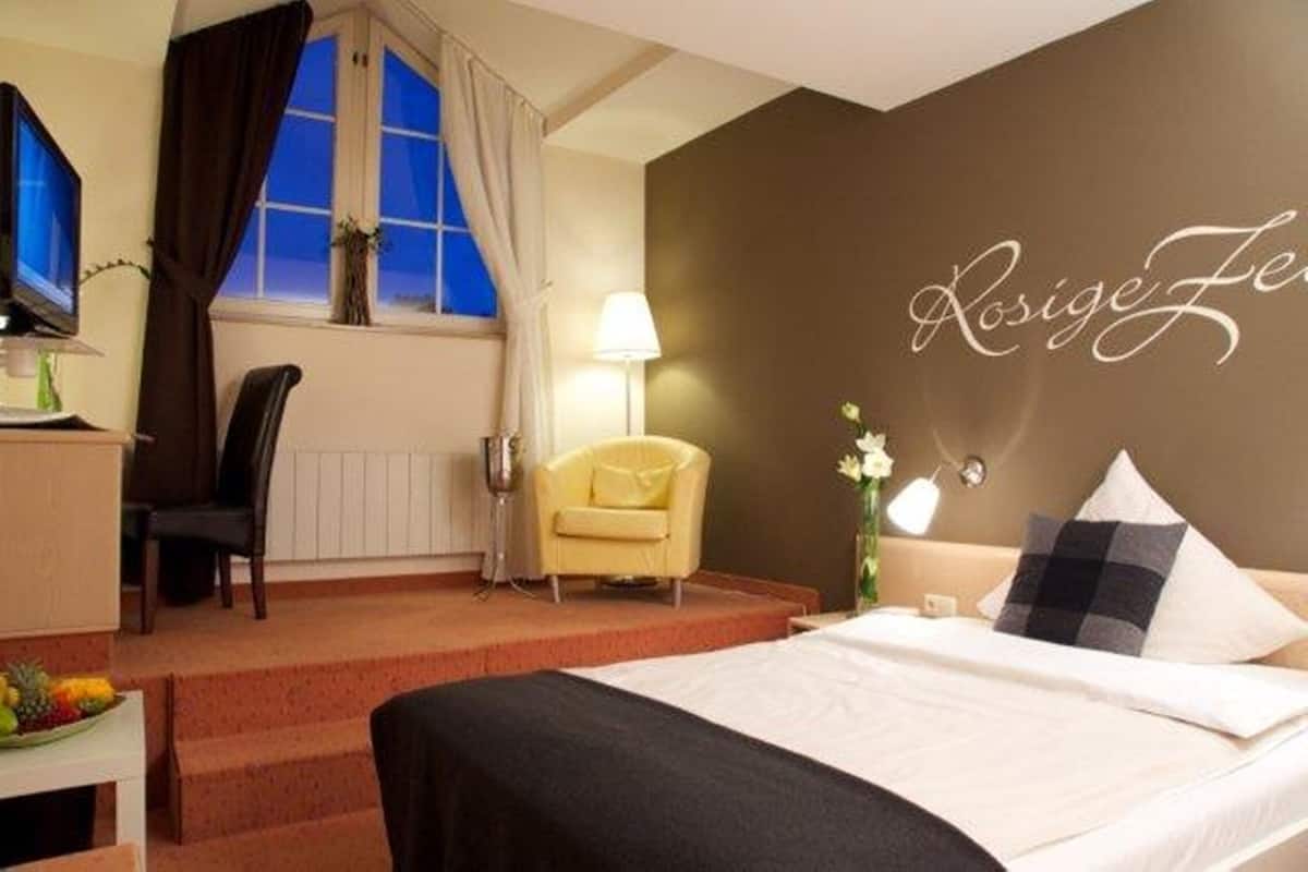 A hotel room with a bed, a desk with a computer, a chair, a lamp, and a window with curtains.