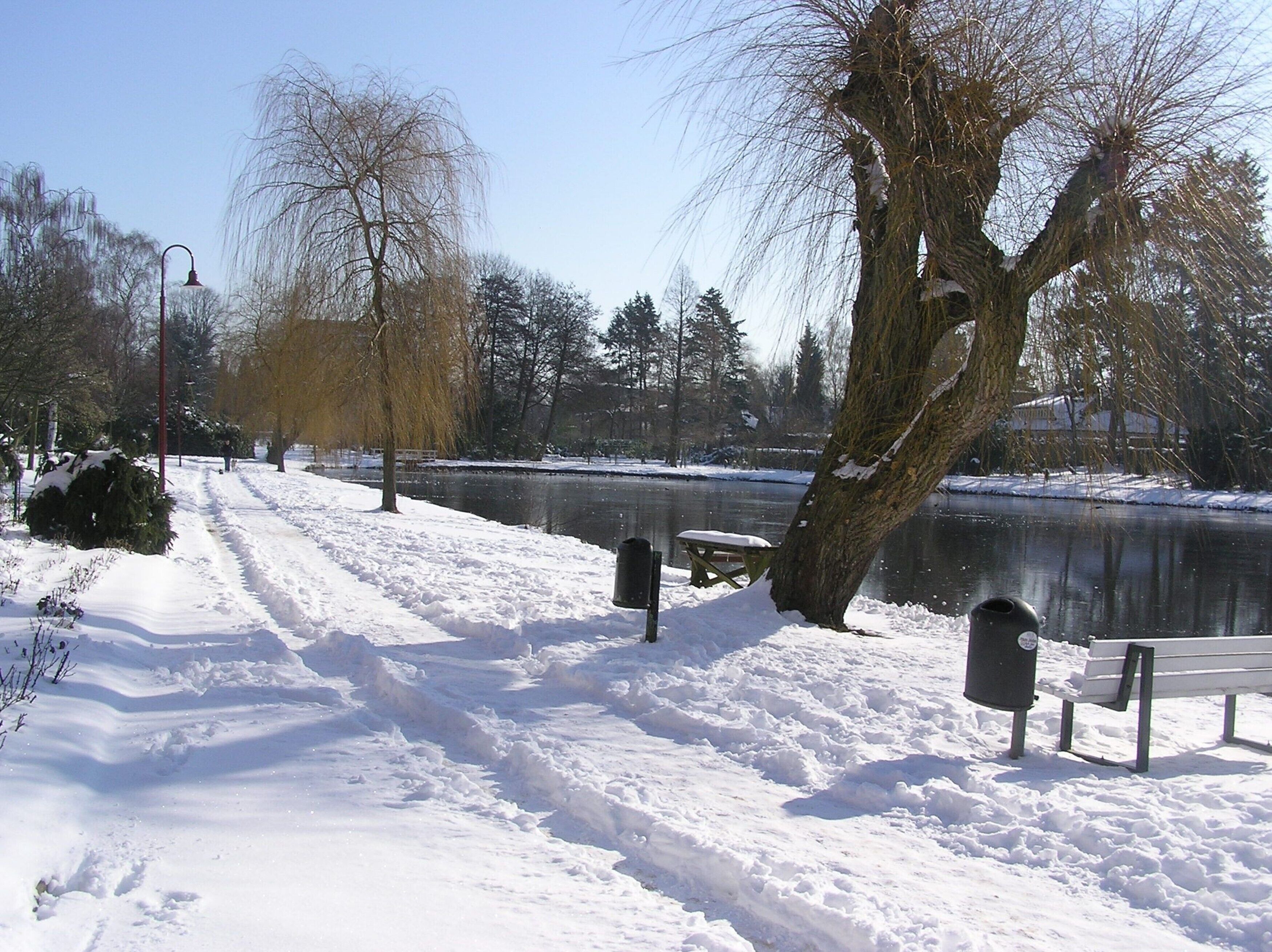A snow-covered park with a bench and trash can near a lake.