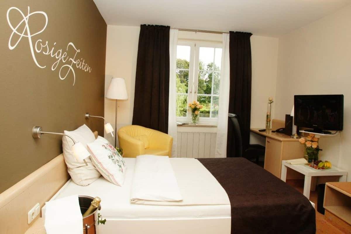 A hotel room with a bed, a desk with a television, a window with curtains, and a wall with a name.