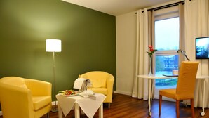 Habitació amb paret verda, sòl de fusta, butaques grogues, taula petita amb estovatge blanc, làmpada de peu, televisió i finestra amb cortines.