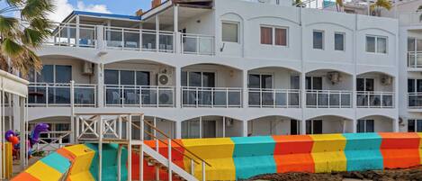 A multi-story hotel with balconies, a colorful slide, and a sandy beach area.