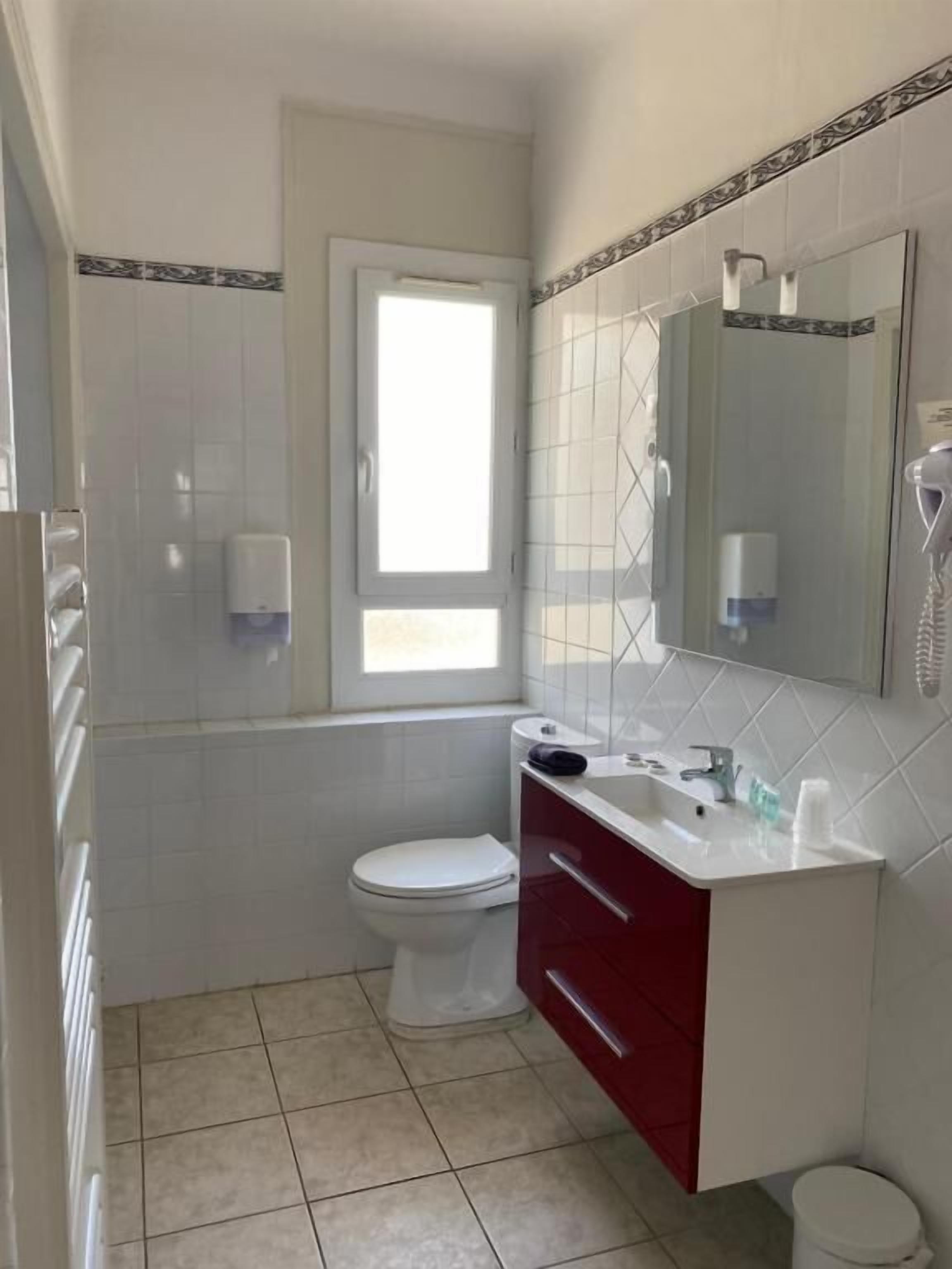 Un baño con inodoro blanco, tocador rojo, un espejo y una ventana.
