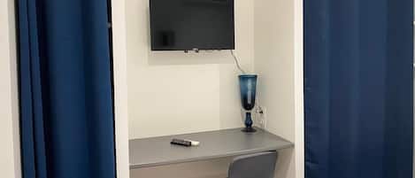 A small room with a desk, a chair, a television mounted on the wall, and a blue curtain.