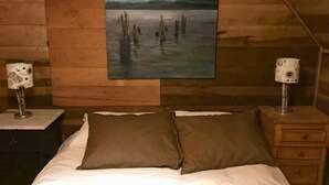Ein Schlafzimmer mit Holzbalkendecke, einem Gemälde mit Wassermotiv, zwei Nachttischlampen und einem Bett mit zwei Kissen.