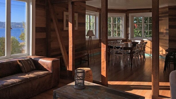 Ein rustikales Blockhaus-Interieur mit Holzboden, Ledersofa, Esstisch mit Stühlen und großen Fenstern mit Blick ins Freie.